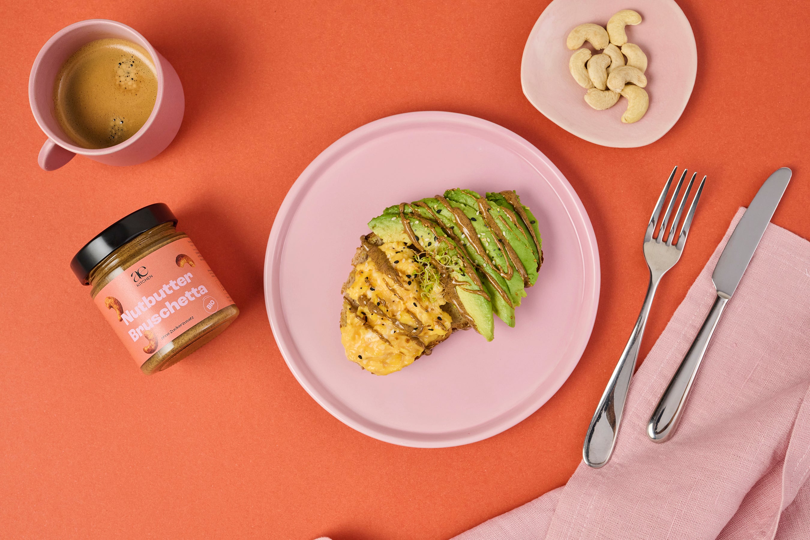 Draufsicht auf Avocado-Toast mit Nutbutter Bruschetta von ae kitchen auf rosa Teller, daneben Kaffee, Cashews und Besteck auf orangem Untergrund