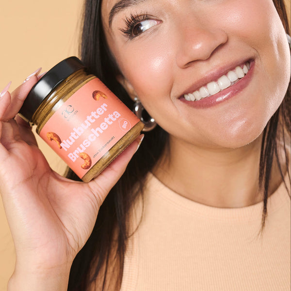 Nahaufnahme einer Frau, die ein Glas Nutbutter Bruschetta von ae kitchen in den Händen hält, vor beigem Hintergrund