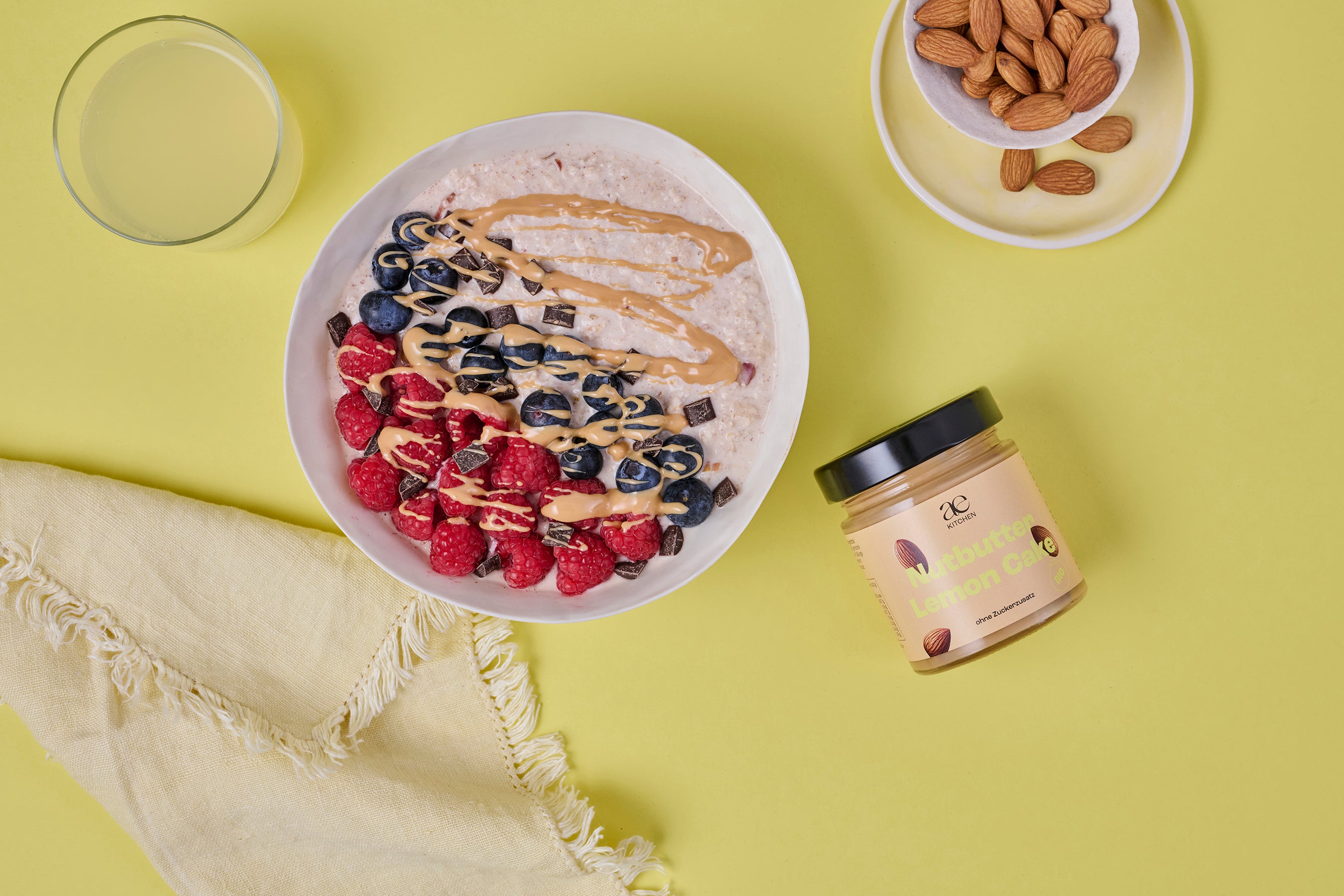 Draufsicht auf eine Porridge-Bowl mit Himbeeren, Blaubeeren, Schokoladenstückchen und Nutbutter Lemon Cake von ae kitchen als Topping, daneben ein Glas Wasser, Mandeln und ein gelbes Tuch auf gelbem Untergrund