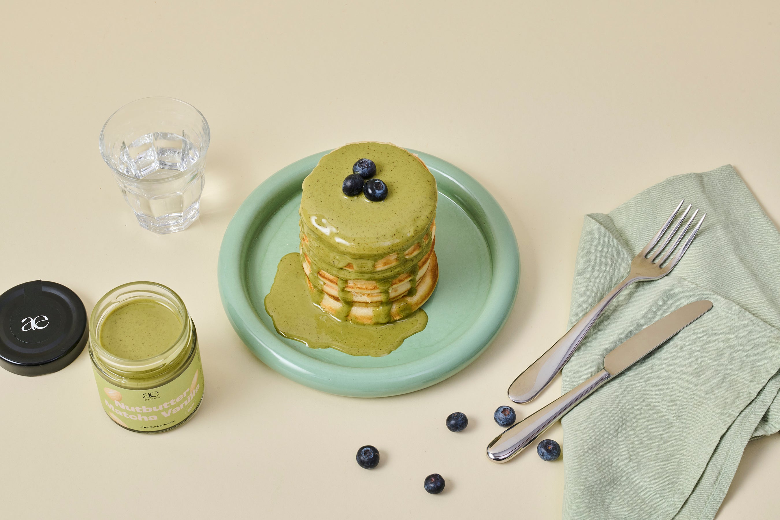 Stapel Pancakes mit Nutbutter Matcha Vanilla von ae kitchen als Topping und frischen Blaubeeren auf grünem Teller, daneben offenes Glas und Besteck