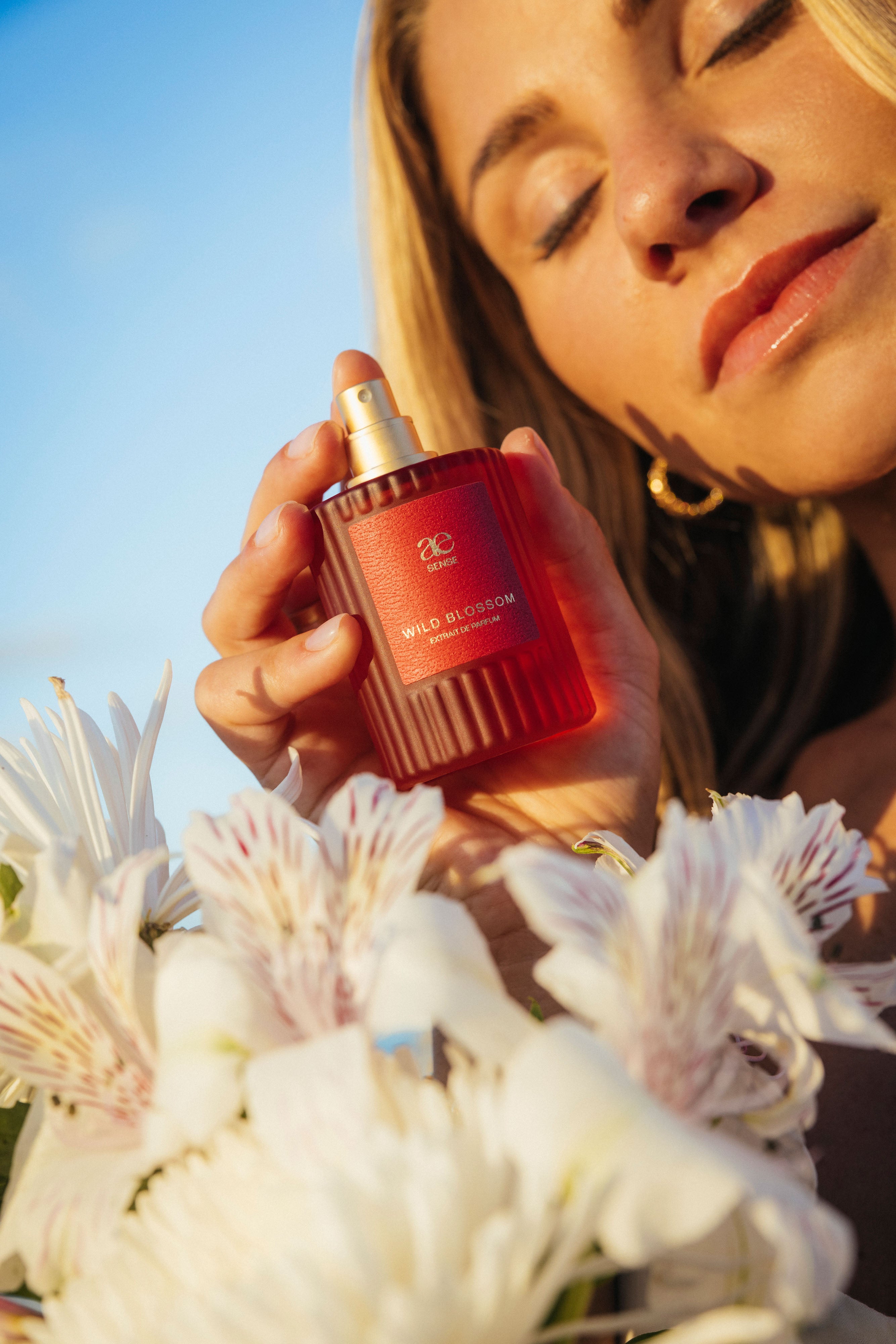 Antonia Elena hält ein Parfümflakon namens Wild Blossom von antonia elena nah an ihrem Gesicht, umgeben von weißen Blüten im Vordergrund. Ihre Augen sind geschlossen und das warme Sonnenlicht fällt auf ihre Haut.