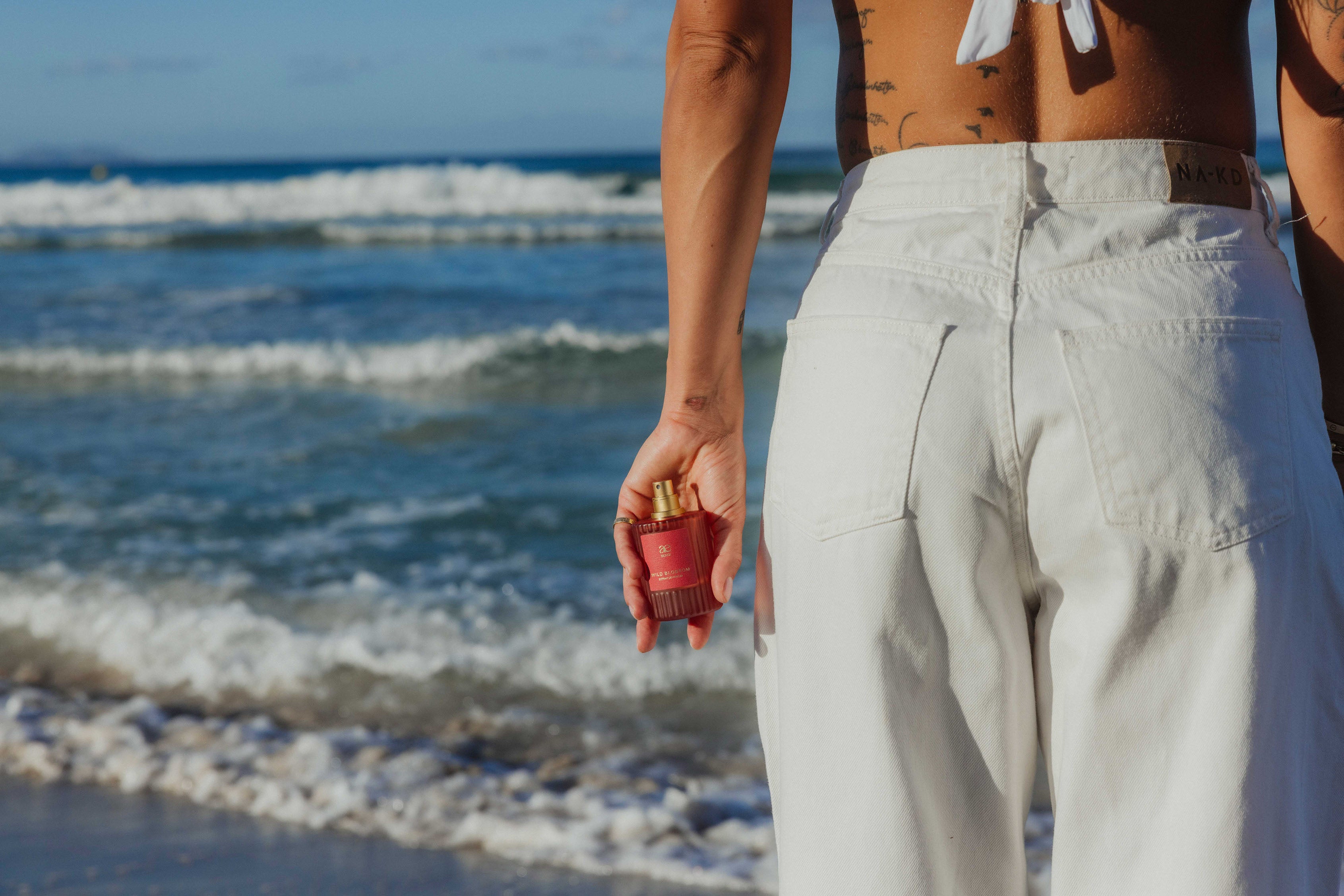 Eine Person steht mit dem Rücken zur Kamera am Strand, trägt weiße Jeans und hält einen roten Parfumflakon in der Hand. Im Hintergrund rollen Wellen ans Ufer.
