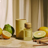 Luminous Glow mit pastellgelber Verpackung, Zitrusfrüchten und Sandelholz auf warmem, mediterranem Hintergrund.
