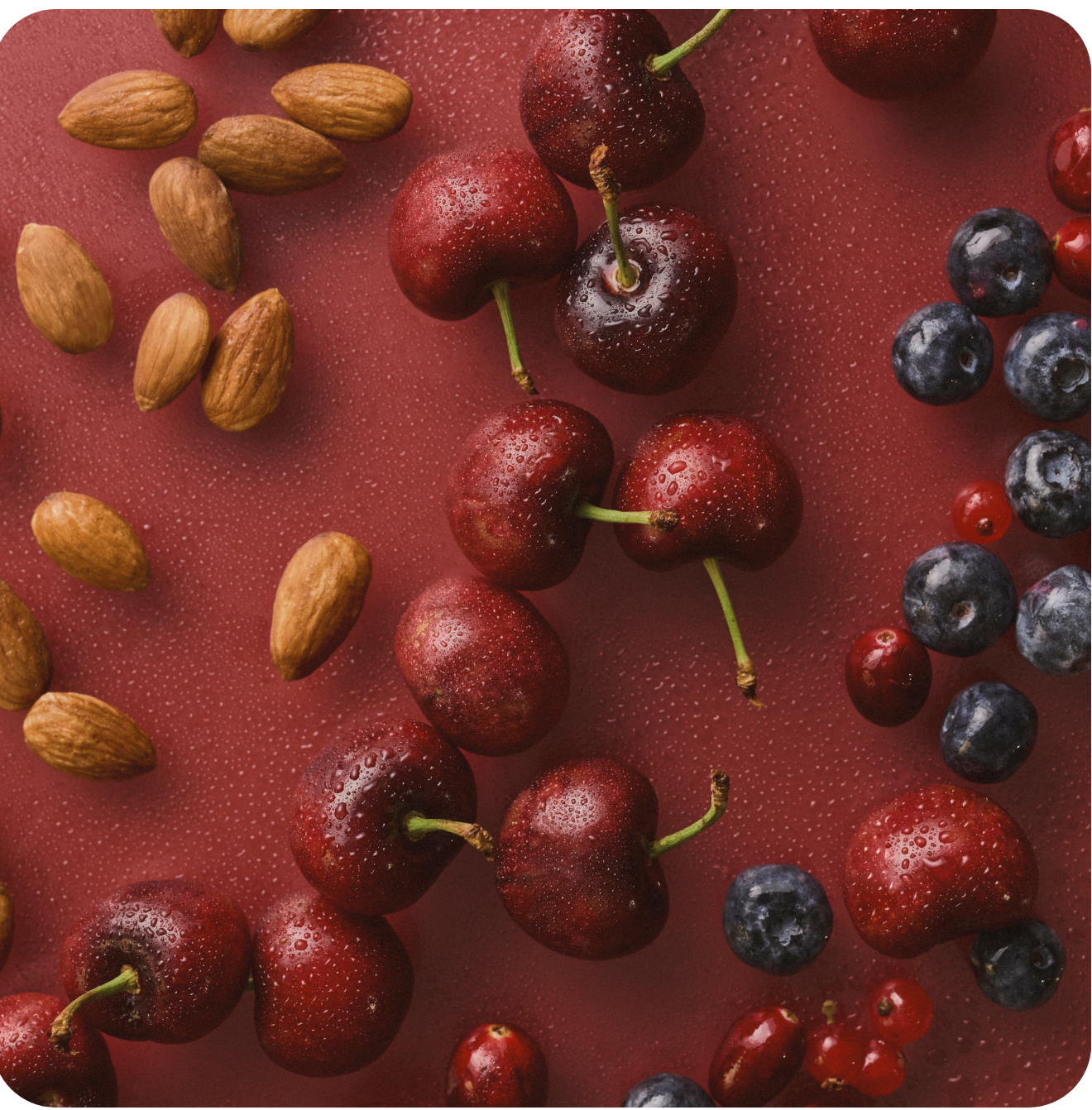 Roter Hintergrund und darauf liege Kirschen, Mandeln, Blaubeeren und Joahnnisbeeren. Es sind einige Wassertropfen auf den Produkten.