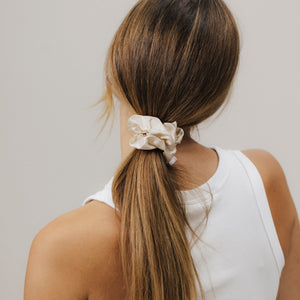 Rückansicht einer Frau mit glatten, schulterlangen Haaren, die zu einem niedrigen Pferdeschwanz gebunden sind. Der Zopf ist mit einem beigefarbenen Seiden-Scrunchie fixiert. Sie trägt ein weißes ärmelloses Oberteil.