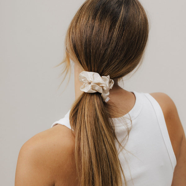 Rückansicht einer Frau mit glatten, schulterlangen Haaren, die zu einem niedrigen Pferdeschwanz gebunden sind. Der Zopf ist mit einem beigefarbenen Seiden-Scrunchie fixiert. Sie trägt ein weißes ärmelloses Oberteil.